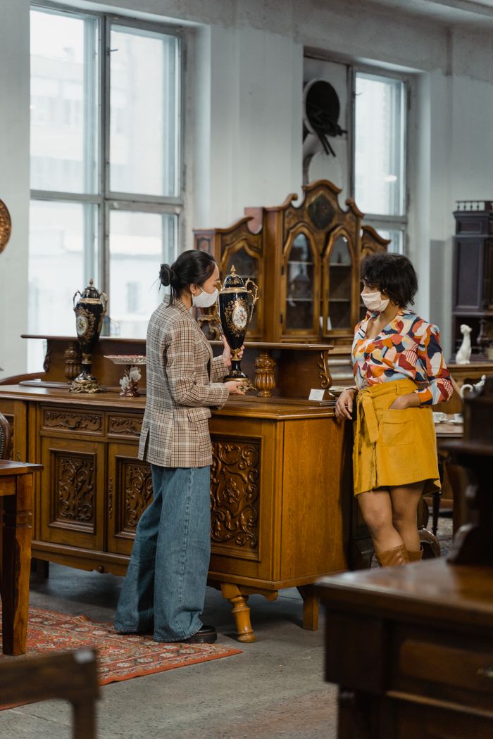 Two women wearing face masks browsing furniture in a vintage shop indoors.